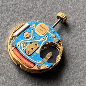 May include: A close-up of a Zenith watch movement, featuring a blue circuit board with gold-colored components. The watch movement has a gold-colored metal casing, a red coil, and a gold-colored crown. The text "ZENITH" and "SWISS V8" are visible.