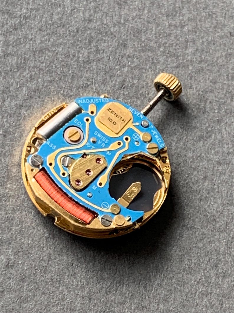 May include: A close-up of a Zenith watch movement, featuring a blue circuit board with gold-colored components. The watch movement has a gold-colored metal casing, a red coil, and a gold-colored crown. The text "ZENITH" and "SWISS V8" are visible.