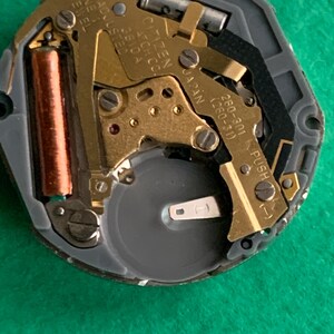 May include: Close-up of a Citizen watch movement. The mechanism features gold-colored gears, a copper-colored coil, and a silver battery. Text on the movement reads "CITIZEN WATCH CO. JAPAN" and "UNADJUSTED ONE JEWEL".