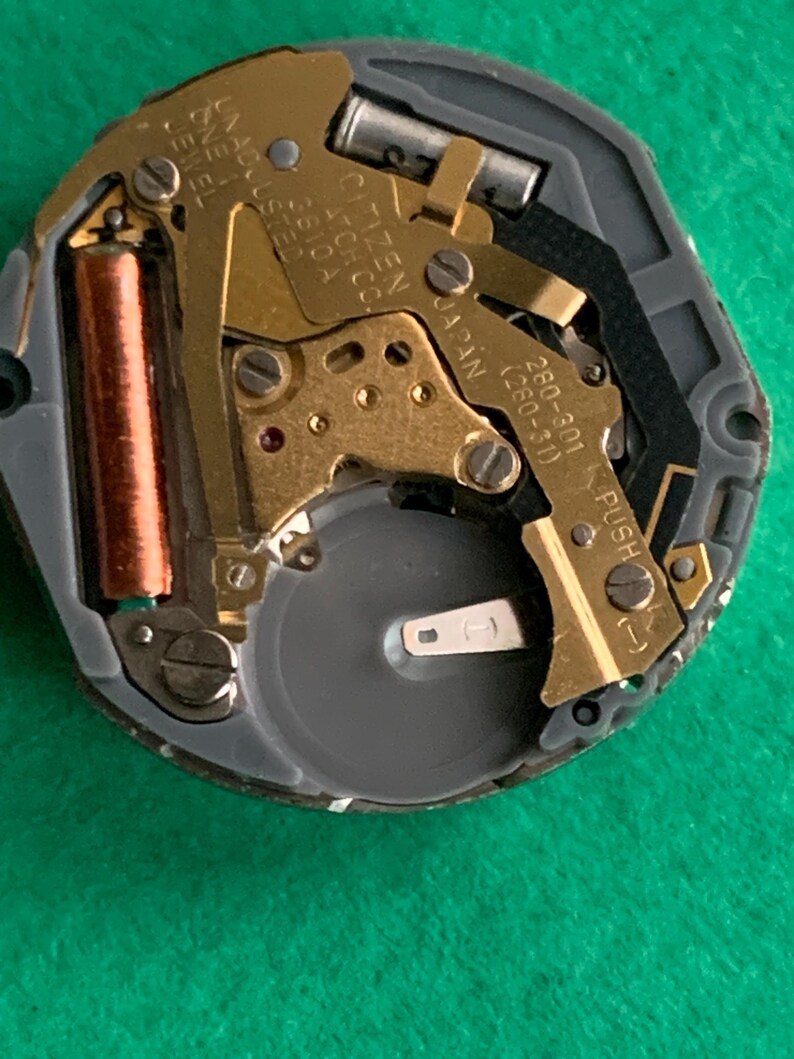 May include: Close-up of a Citizen watch movement. The mechanism features gold-colored gears, a copper-colored coil, and a silver battery. Text on the movement reads "CITIZEN WATCH CO. JAPAN" and "UNADJUSTED ONE JEWEL".