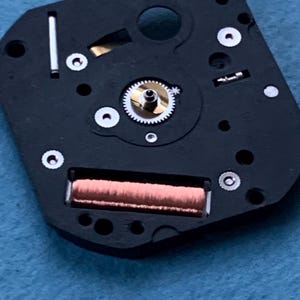 May include: Close-up of a watch movement. The black base features a copper coil, a small gear, and several silver-colored screws. The intricate design showcases the internal components of a timepiece.