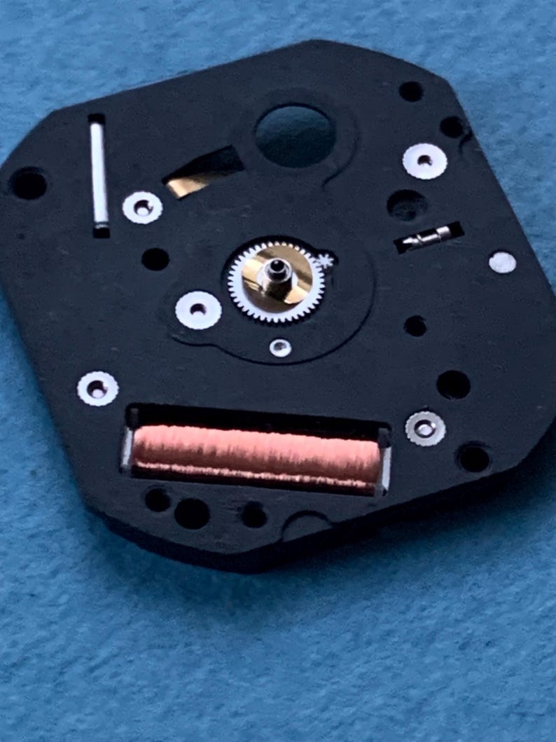May include: Close-up of a watch movement. The black base features a copper coil, a small gear, and several silver-colored screws. The intricate design showcases the internal components of a timepiece.