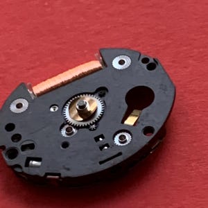 May include: Close-up of a watch movement component. The black, irregularly shaped piece features a small gear, a copper coil, and several silver-colored screws. The background is a solid red color.