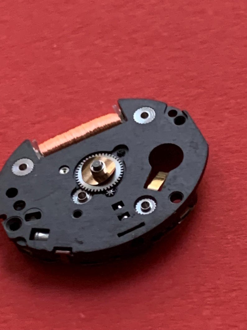 May include: Close-up of a watch movement component. The black, irregularly shaped piece features a small gear, a copper coil, and several silver-colored screws. The background is a solid red color.