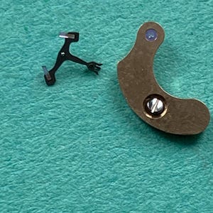 May include: Close-up of two watch parts. One is a small, black, multi-pronged lever. The other is a curved, bronze-colored piece with a screw and a small, purple-colored jewel. The background is a teal-colored surface.