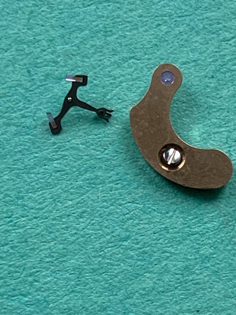 May include: Close-up of two watch parts. One is a small, black, multi-pronged lever. The other is a curved, bronze-colored piece with a screw and a small, purple-colored jewel. The background is a teal-colored surface.