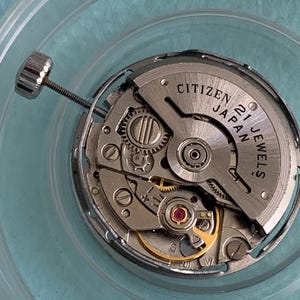 May include: Close-up of a Citizen Japan 21 Jewels watch movement. The silver-toned mechanism features intricate gears, a balance wheel, and a crown stem. The watch movement is set against a light blue background.