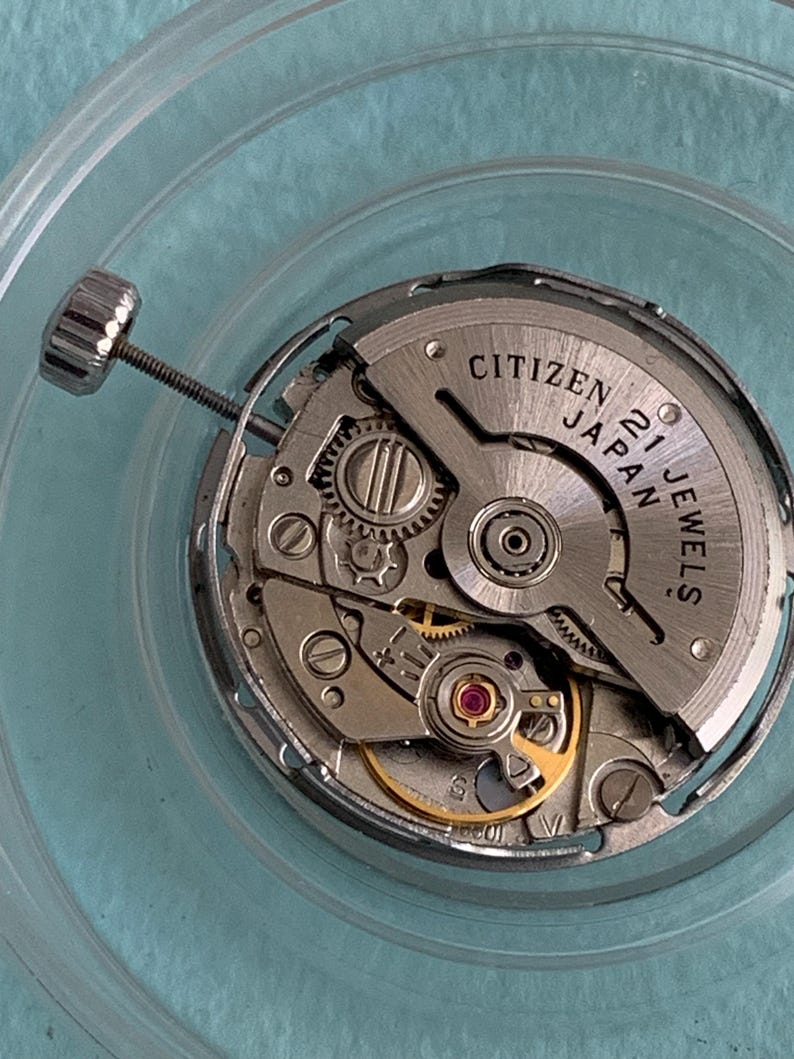 May include: Close-up of a Citizen Japan 21 Jewels watch movement. The silver-toned mechanism features intricate gears, a balance wheel, and a crown stem. The watch movement is set against a light blue background.