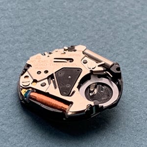 K&ouml;nnte beinhalten: Nahaufnahme eines Seiko-Uhrwerks. Der Uhrenmechanismus verf&uuml;gt &uuml;ber eine Kupferspule, verschiedene Zahnr&auml;der und den Text "JAPAN" und "SEIKO TIME CORP." auf den Metallkomponenten. Das Uhrwerk befindet sich auf einer blauen Oberfl&auml;che.