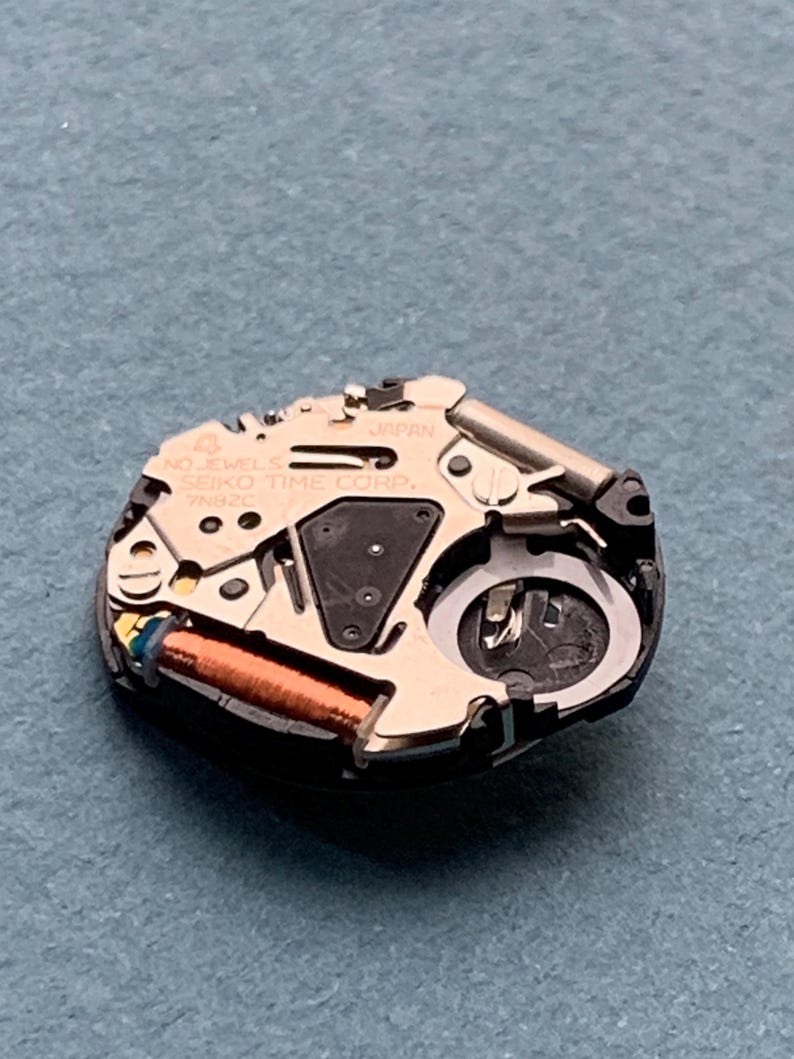 K&ouml;nnte beinhalten: Nahaufnahme eines Seiko-Uhrwerks. Der Uhrenmechanismus verf&uuml;gt &uuml;ber eine Kupferspule, verschiedene Zahnr&auml;der und den Text "JAPAN" und "SEIKO TIME CORP." auf den Metallkomponenten. Das Uhrwerk befindet sich auf einer blauen Oberfl&auml;che.