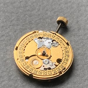 May include: A gold-toned watch movement with intricate gears and a date dial. The dial features numbers from 1 to 31, indicating the days of the month. A small, threaded crown is visible at the top.