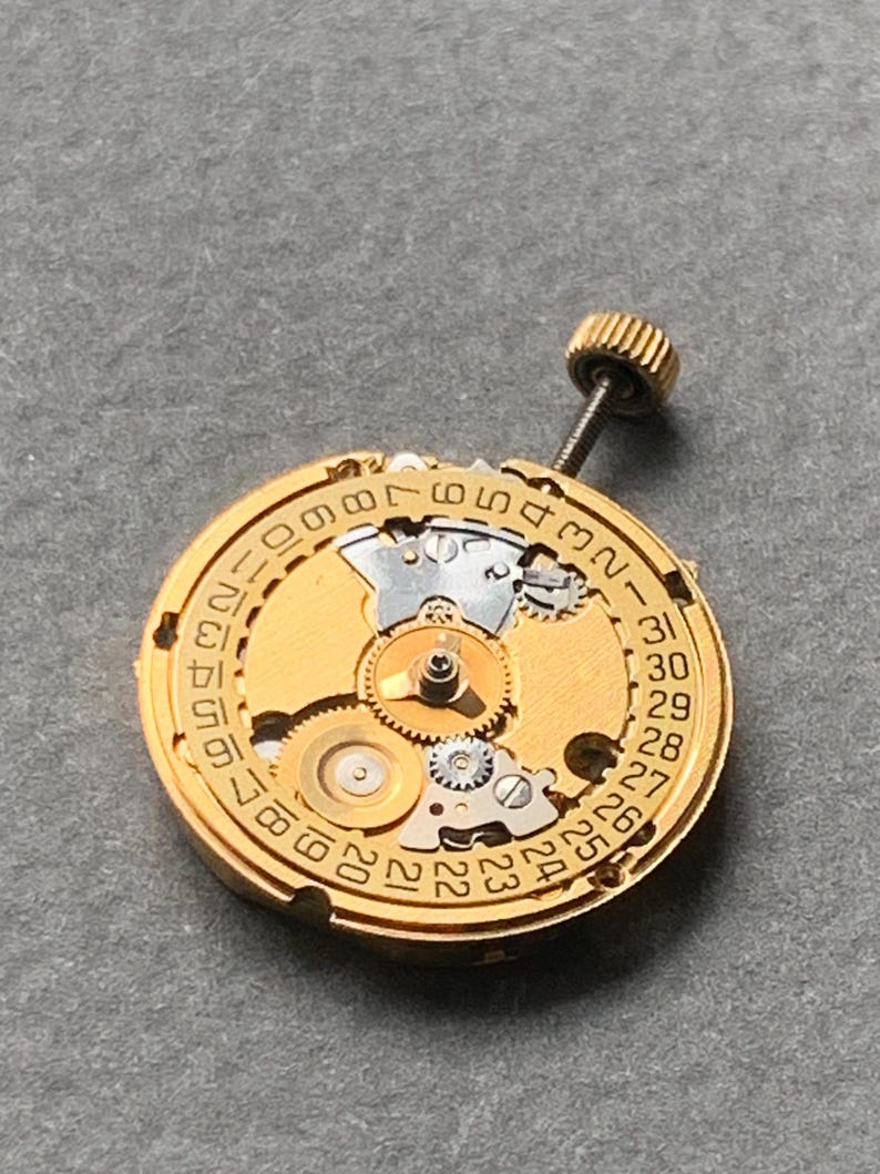 May include: A gold-toned watch movement with intricate gears and a date dial. The dial features numbers from 1 to 31, indicating the days of the month. A small, threaded crown is visible at the top.