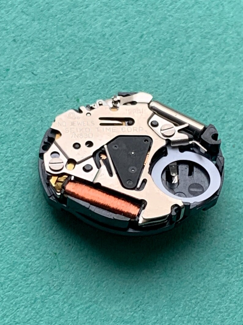 May include: Close-up of a watch movement with the text "NO JEWELS SEIKO TIME CORP 7N83D". The movement features metallic components, a copper coil, and a black plastic base, set against a teal background.