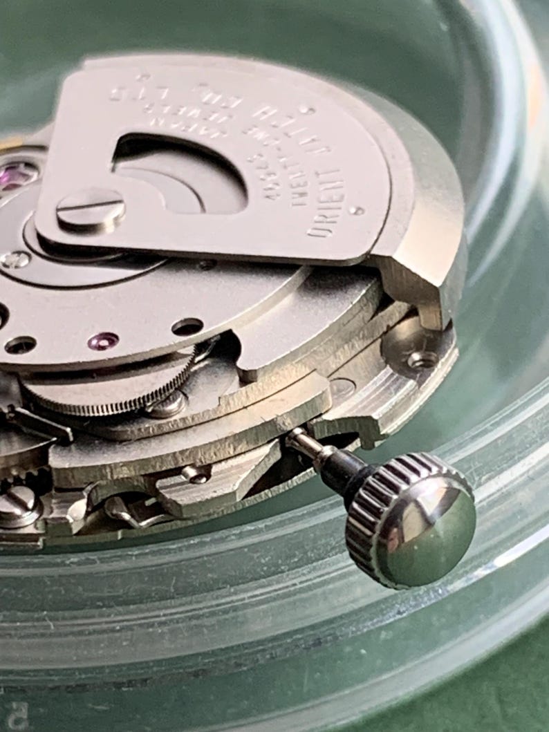 May include: Detailed view of a mechanical watch movement, revealing complex gears and mechanisms. The silver metal components are finely crafted, with a visible winding crown. The watch is marked with "ORIENT" and "JAPAN".
