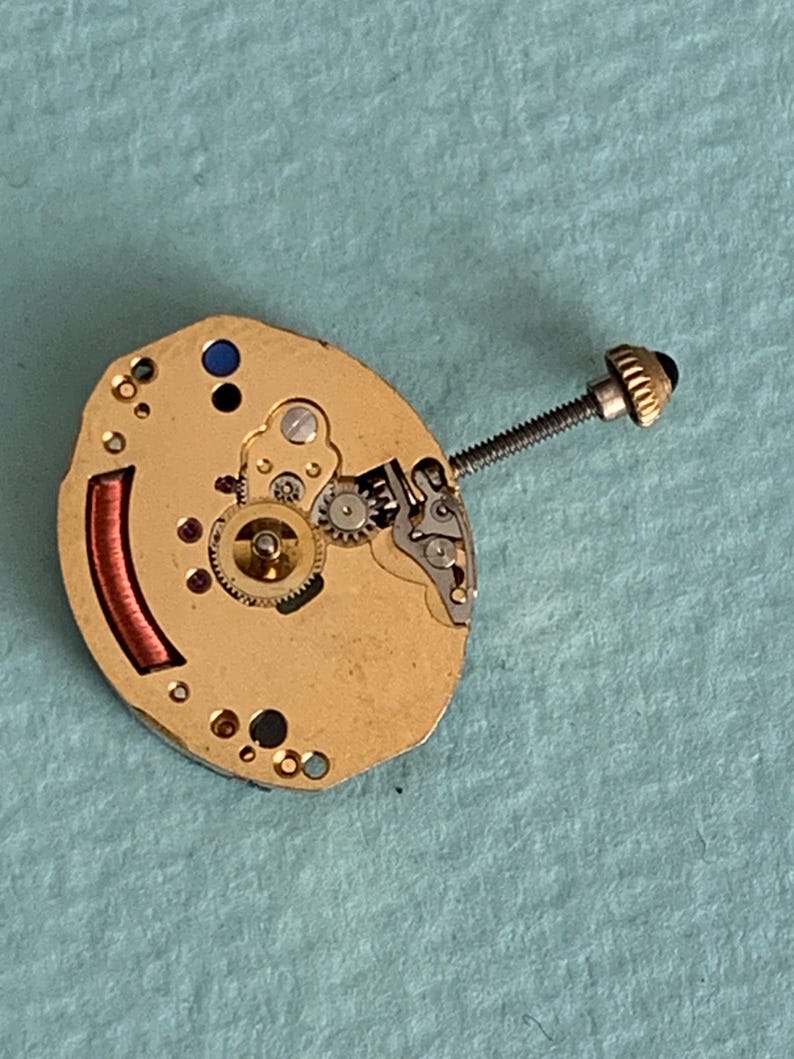May include: Close-up of a watch movement with gold-colored metal components. The movement features small gears, a red coil, and a screw with a black head. The background is a light blue surface.