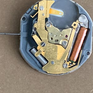 May include: A close-up of a watch movement with gold-colored metal components, a gray plastic base, and a copper-colored coil. The text "CITIZEN WATCH CO. UNADJUSTED 38014 ONE 1 JEWEL" is visible.