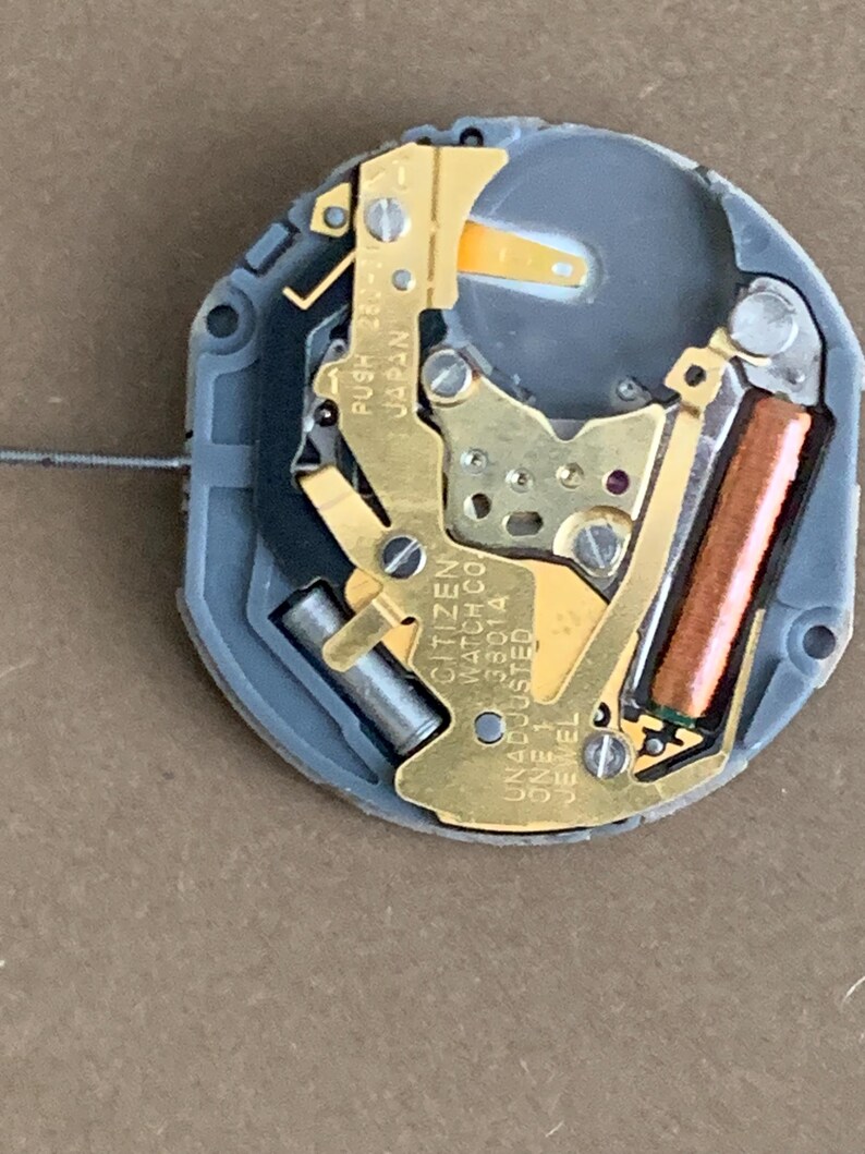 May include: A close-up of a watch movement with gold-colored metal components, a gray plastic base, and a copper-colored coil. The text "CITIZEN WATCH CO. UNADJUSTED 38014 ONE 1 JEWEL" is visible.