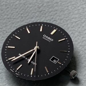 Puede incluir: Una esfera de reloj Casio Quartz negra con marcadores de hora y agujas doradas. La esfera tiene las palabras "CASIO QUARTZ" y "WATER RESIST" impresas. Tambi&eacute;n tiene una ventana de fecha y una corona plateada.