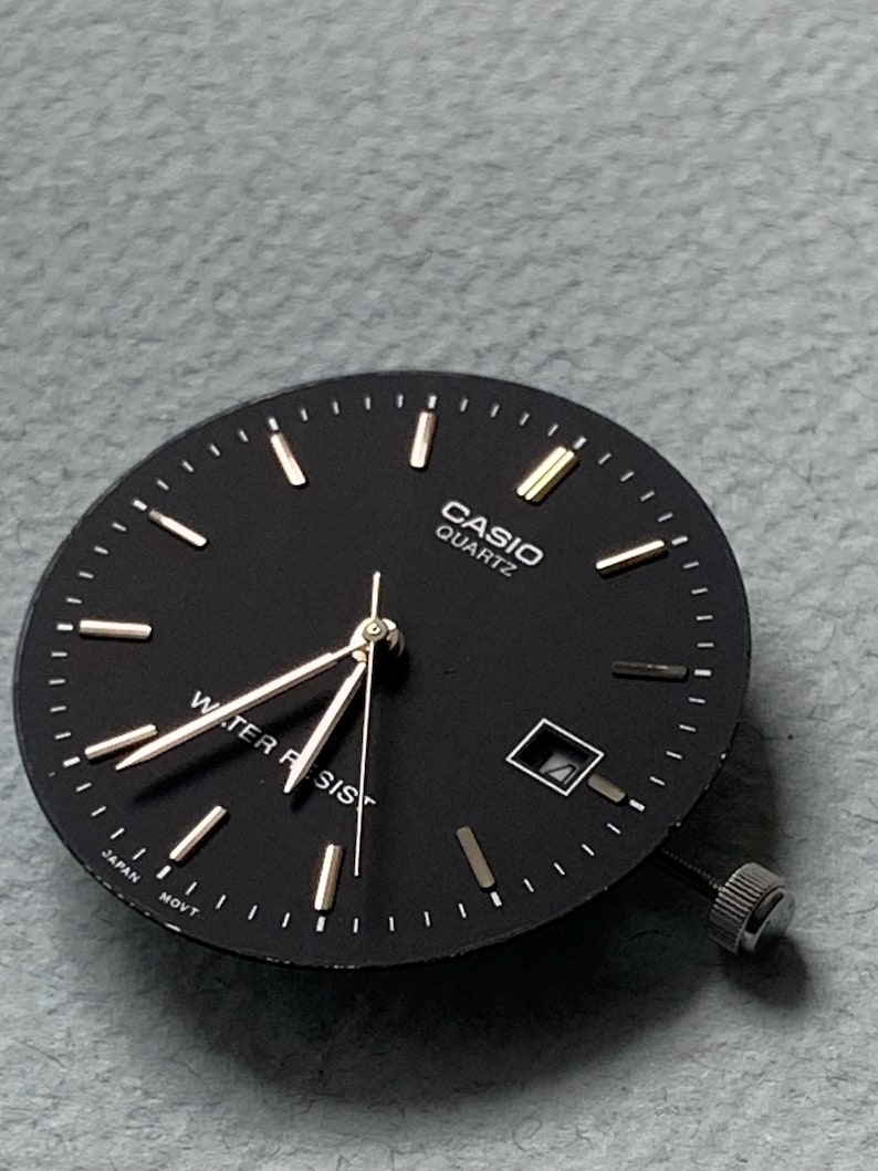 Puede incluir: Una esfera de reloj Casio Quartz negra con marcadores de hora y agujas doradas. La esfera tiene las palabras "CASIO QUARTZ" y "WATER RESIST" impresas. Tambi&eacute;n tiene una ventana de fecha y una corona plateada.