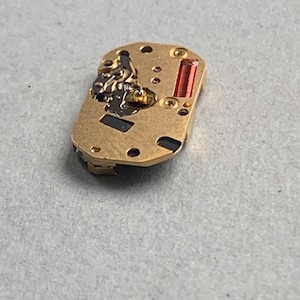 May include: A small, rectangular watch movement with a gold-colored metal base. It features various components, including a red rectangular piece and several small screws. The watch movement is designed for use in a timepiece.