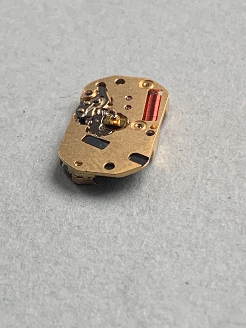 May include: A small, rectangular watch movement with a gold-colored metal base. It features various components, including a red rectangular piece and several small screws. The watch movement is designed for use in a timepiece.