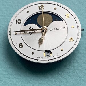 Op de afbeelding: Een witte quartz wijzerplaat met gouden wijzers en accenten. De wijzerplaat heeft een maanfase-indicator, het woord "Royal" en de datum "19". De wijzerplaat is gemarkeerd met "Swiss Made".