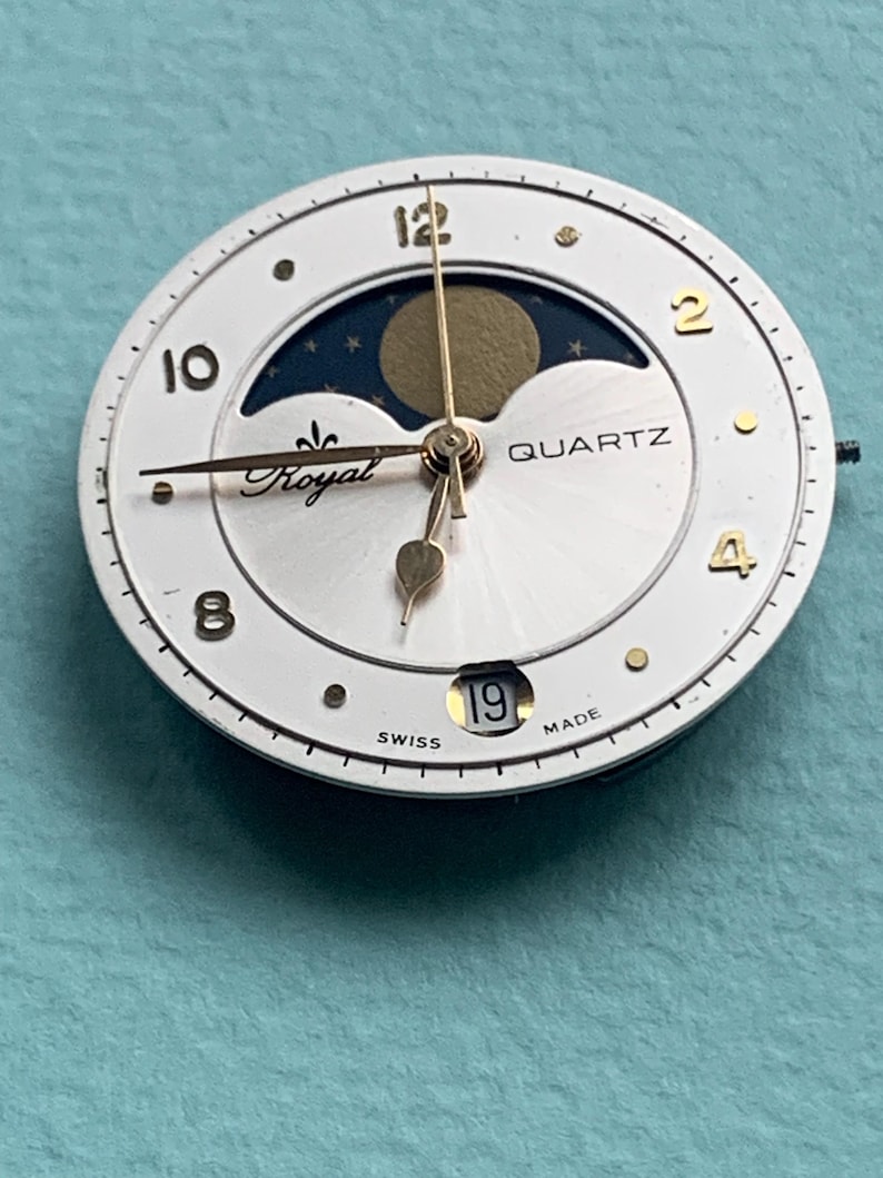 Op de afbeelding: Een witte quartz wijzerplaat met gouden wijzers en accenten. De wijzerplaat heeft een maanfase-indicator, het woord "Royal" en de datum "19". De wijzerplaat is gemarkeerd met "Swiss Made".