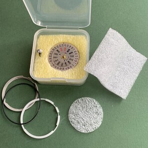 May include: A watch movement repair kit. The clear plastic case contains a silver dial with day and date indicators. Includes black and white rings, a small screw, and a white foam pad. The background is green.