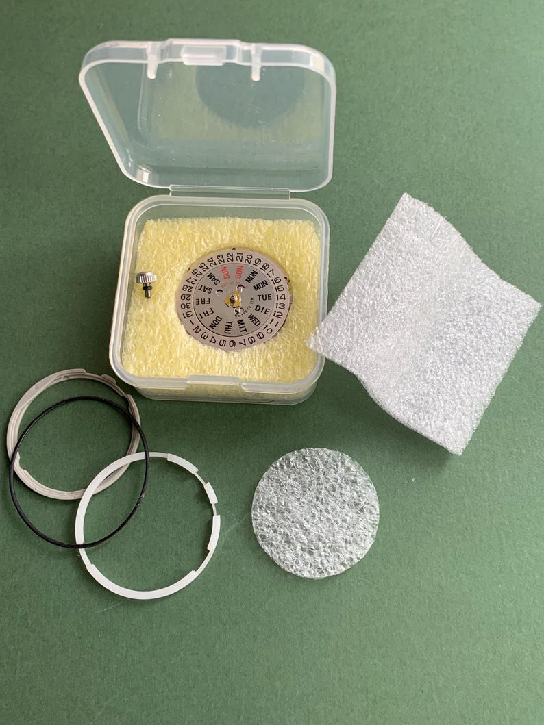 May include: A watch movement repair kit. The clear plastic case contains a silver dial with day and date indicators. Includes black and white rings, a small screw, and a white foam pad. The background is green.