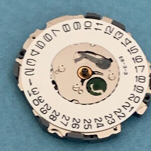 May include: A watch movement component with a white dial featuring black numbers 1-31, indicating the days of the month. The component has a silver-colored inner mechanism with a small green element and the text "E6-3-3".