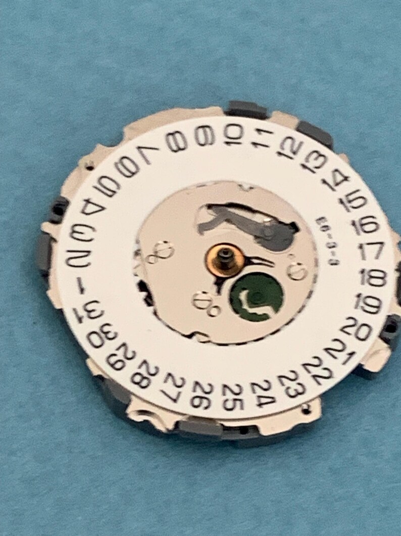 May include: A watch movement component with a white dial featuring black numbers 1-31, indicating the days of the month. The component has a silver-colored inner mechanism with a small green element and the text "E6-3-3".
