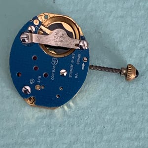 May include: A close-up of a watch movement with a blue circuit board and gold-colored components. The movement has a screw and a small crown. The text "SWISS SIX 6 JEWELS" is visible.