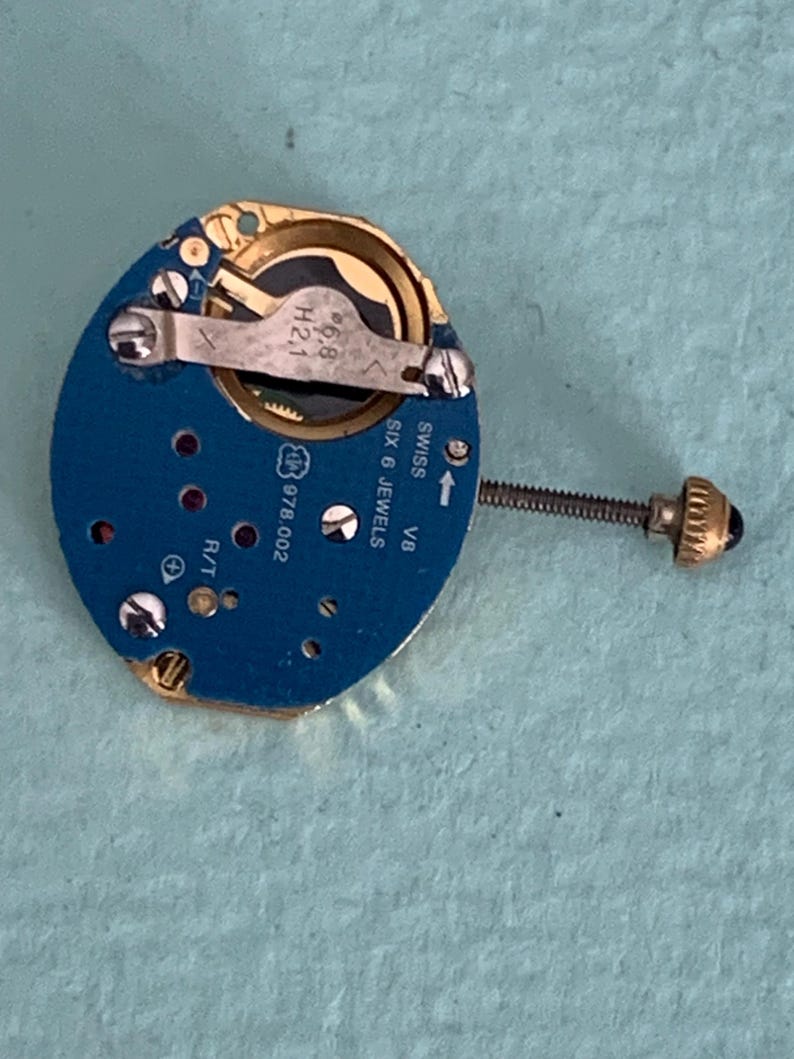 May include: A close-up of a watch movement with a blue circuit board and gold-colored components. The movement has a screw and a small crown. The text "SWISS SIX 6 JEWELS" is visible.