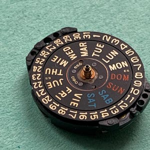 May include: A close-up of a watch movement with a black dial and gold-colored numerals and text. The dial displays the days of the week in English, Spanish, and French, and the dates 1-31.