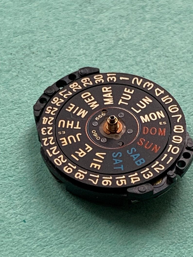 May include: A close-up of a watch movement with a black dial and gold-colored numerals and text. The dial displays the days of the week in English, Spanish, and French, and the dates 1-31.