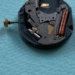 K&ouml;nnte beinhalten: Nahaufnahme eines Uhrwerks mit komplexen mechanischen Komponenten. Der Mechanismus zeigt eine goldfarbene Krone, eine kupferfarbene Spule und verschiedene silberfarbene Metallteile. Das Uhrwerk ist vor einem hellblauen Hintergrund platziert.