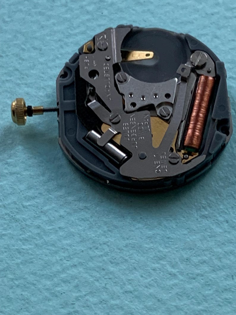 K&ouml;nnte beinhalten: Nahaufnahme eines Uhrwerks mit komplexen mechanischen Komponenten. Der Mechanismus zeigt eine goldfarbene Krone, eine kupferfarbene Spule und verschiedene silberfarbene Metallteile. Das Uhrwerk ist vor einem hellblauen Hintergrund platziert.