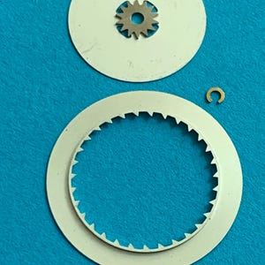 May include: A collection of watch parts, including two white circular components, a small gear, and a gold-colored C-shaped piece. The larger circular piece has a toothed edge. The parts are arranged against a blue background.
