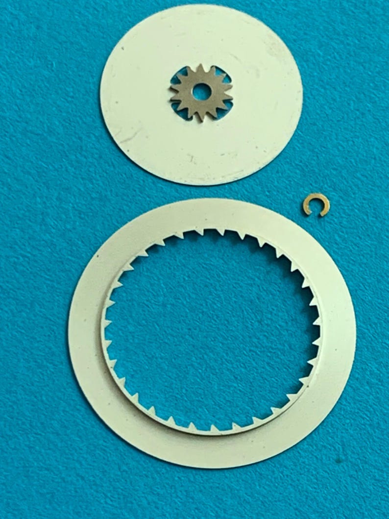 May include: A collection of watch parts, including two white circular components, a small gear, and a gold-colored C-shaped piece. The larger circular piece has a toothed edge. The parts are arranged against a blue background.