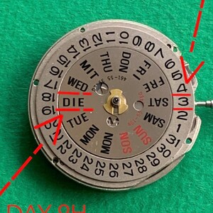 May include: Close-up of a watch movement with a silver dial and black numerals. The dial displays the days of the week and the date. Red lines and text indicate "DATE 3H" and "DAY 9H".
