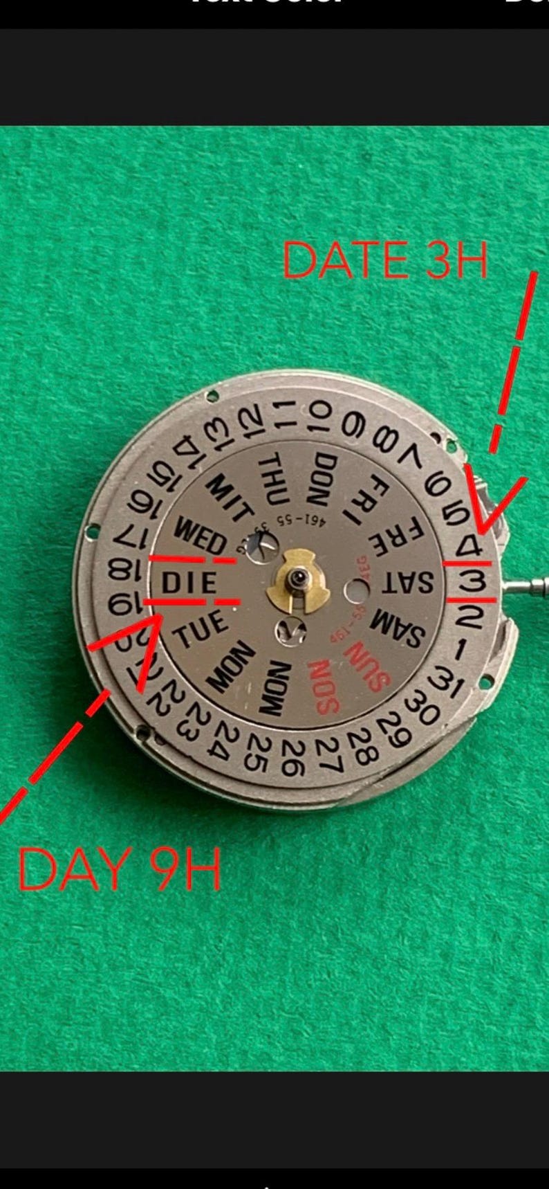 May include: Close-up of a watch movement with a silver dial and black numerals. The dial displays the days of the week and the date. Red lines and text indicate "DATE 3H" and "DAY 9H".