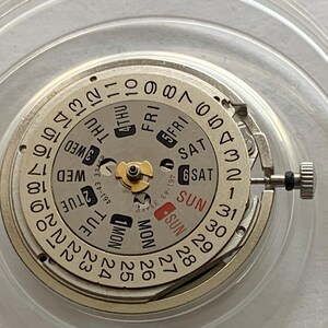 May include: Close-up of a watch movement with a silver-colored dial and a calendar display. The dial features day and date indicators, with the days of the week and numbers clearly visible. The watch mechanism is encased in a clear plastic holder.