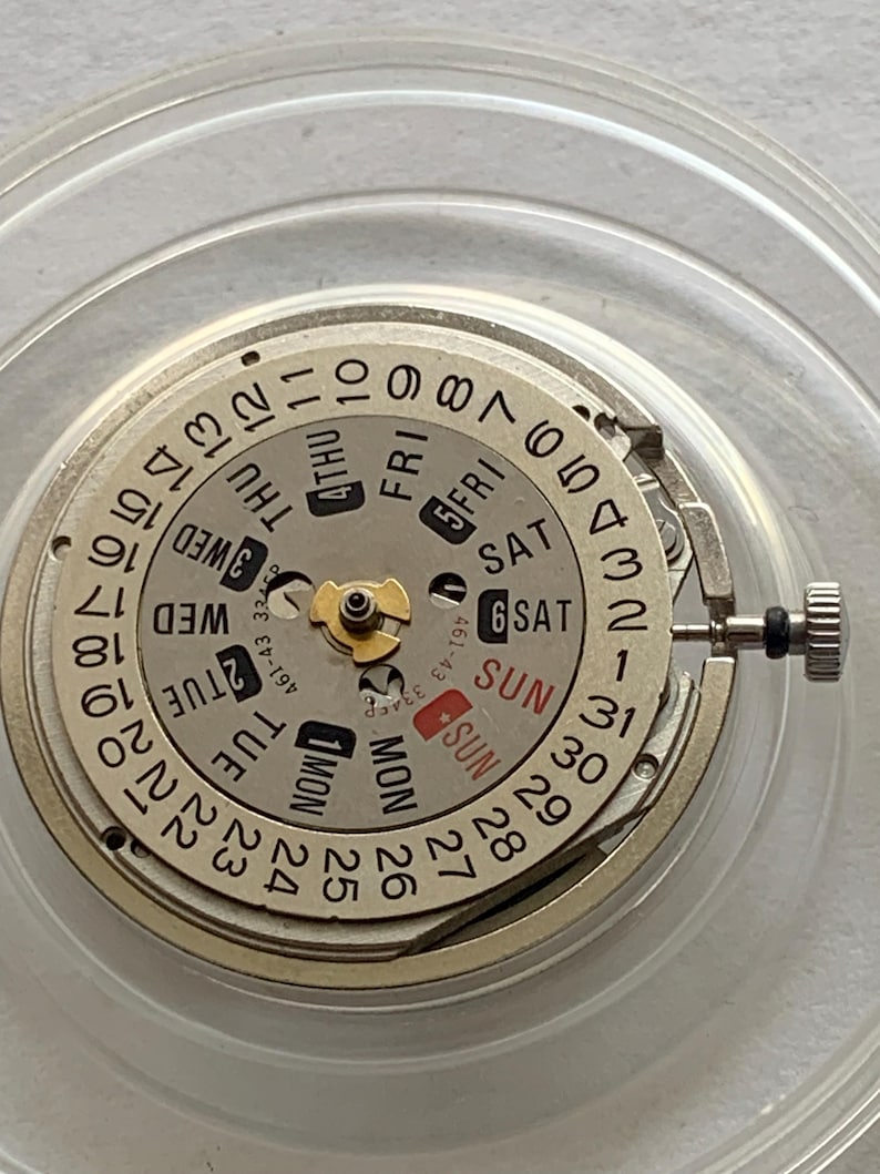 May include: Close-up of a watch movement with a silver-colored dial and a calendar display. The dial features day and date indicators, with the days of the week and numbers clearly visible. The watch mechanism is encased in a clear plastic holder.