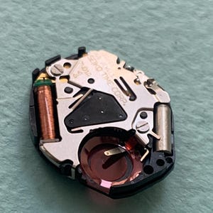 May include: A close-up of a watch movement with the text "SEIKO TIME CORP." and "16 JEWELS" visible. The movement has a black plastic base with silver-coloured metal components, a copper coil, and a red-tinted crystal.