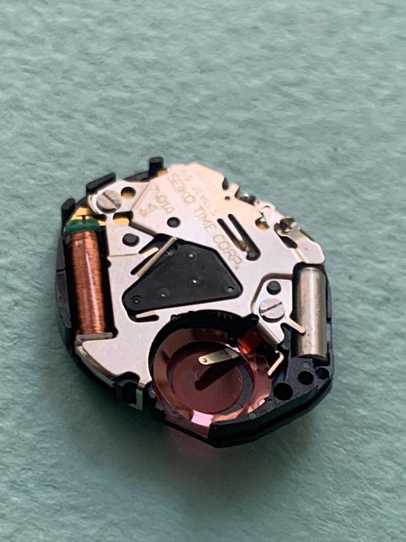 May include: A close-up of a watch movement with the text "SEIKO TIME CORP." and "16 JEWELS" visible. The movement has a black plastic base with silver-coloured metal components, a copper coil, and a red-tinted crystal.