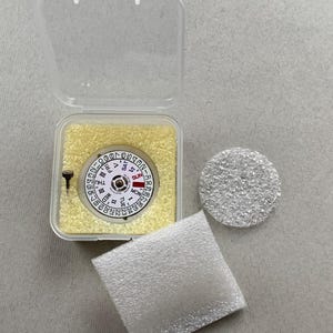May include: A watch movement with a white dial, black numerals, and day/date indicators, displayed in a clear plastic case with yellow padding. Includes a small screw, a round silver component, and a white foam pad.