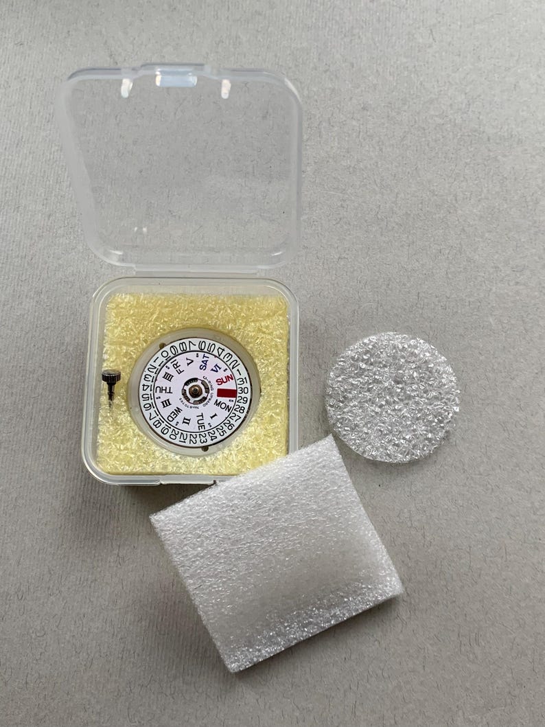 May include: A watch movement with a white dial, black numerals, and day/date indicators, displayed in a clear plastic case with yellow padding. Includes a small screw, a round silver component, and a white foam pad.