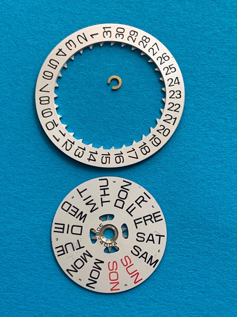 May include: Watch parts, including a silver-colored date wheel with numbers 1-31 and a day wheel with days of the week in black and red. A small gold-colored component is also present. The parts are on a blue background.