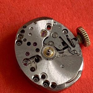 May include: Close-up of a vintage watch movement with intricate gears, jewels, and a gold-colored crown. The silver-toned mechanism is set against a red background, showcasing the detailed craftsmanship of the watch's internal components.