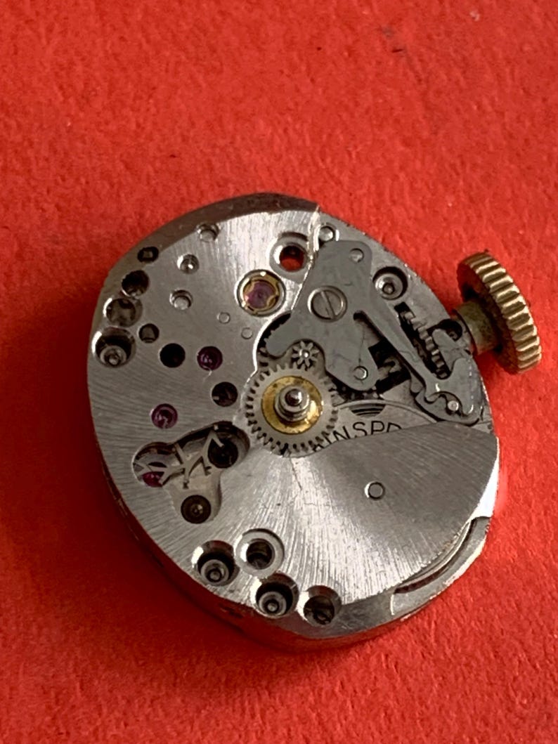 May include: Close-up of a vintage watch movement with intricate gears, jewels, and a gold-colored crown. The silver-toned mechanism is set against a red background, showcasing the detailed craftsmanship of the watch's internal components.